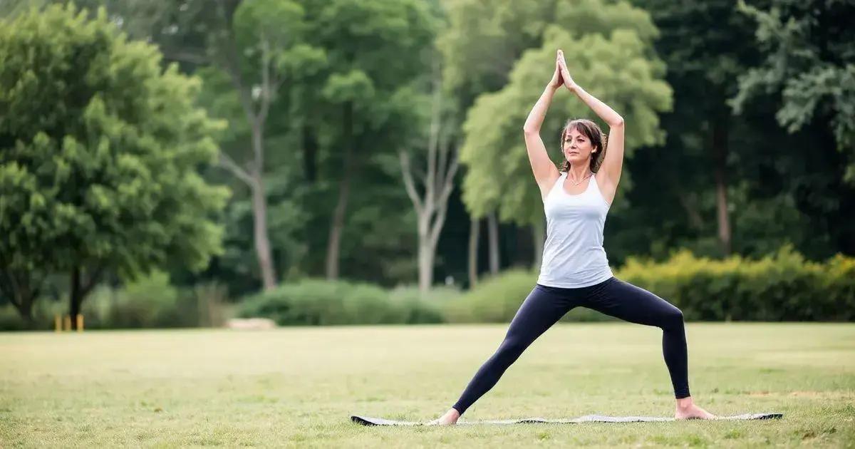 A person is performing a yoga pose outdoors on a grassy field. The individual is standing on a yoga mat with legs spread wide apart and arms raised above the head, palms together. The person is wearing a white tank top and dark leggings. The background features lush green trees and bushes, indicating a natural park or garden setting. The image captures a moment of physical exercise and mindfulness in a serene outdoor environment.