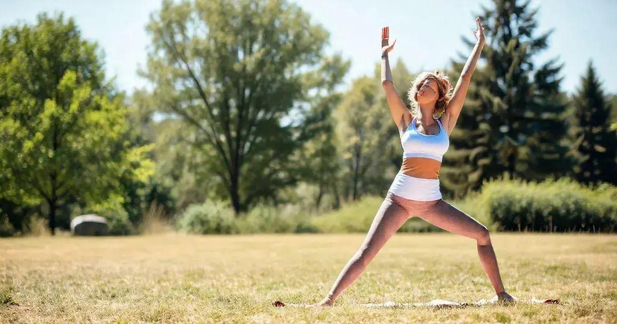 A woman is practicing yoga outdoors on a sunny day. She is standing on a yoga mat in a wide, grassy area surrounded by trees. She is wearing a light blue sports bra and white shorts, and she is performing a yoga pose with her legs spread apart and her arms raised above her head. The background features lush green trees and a clear sky, indicating a peaceful and natural setting.