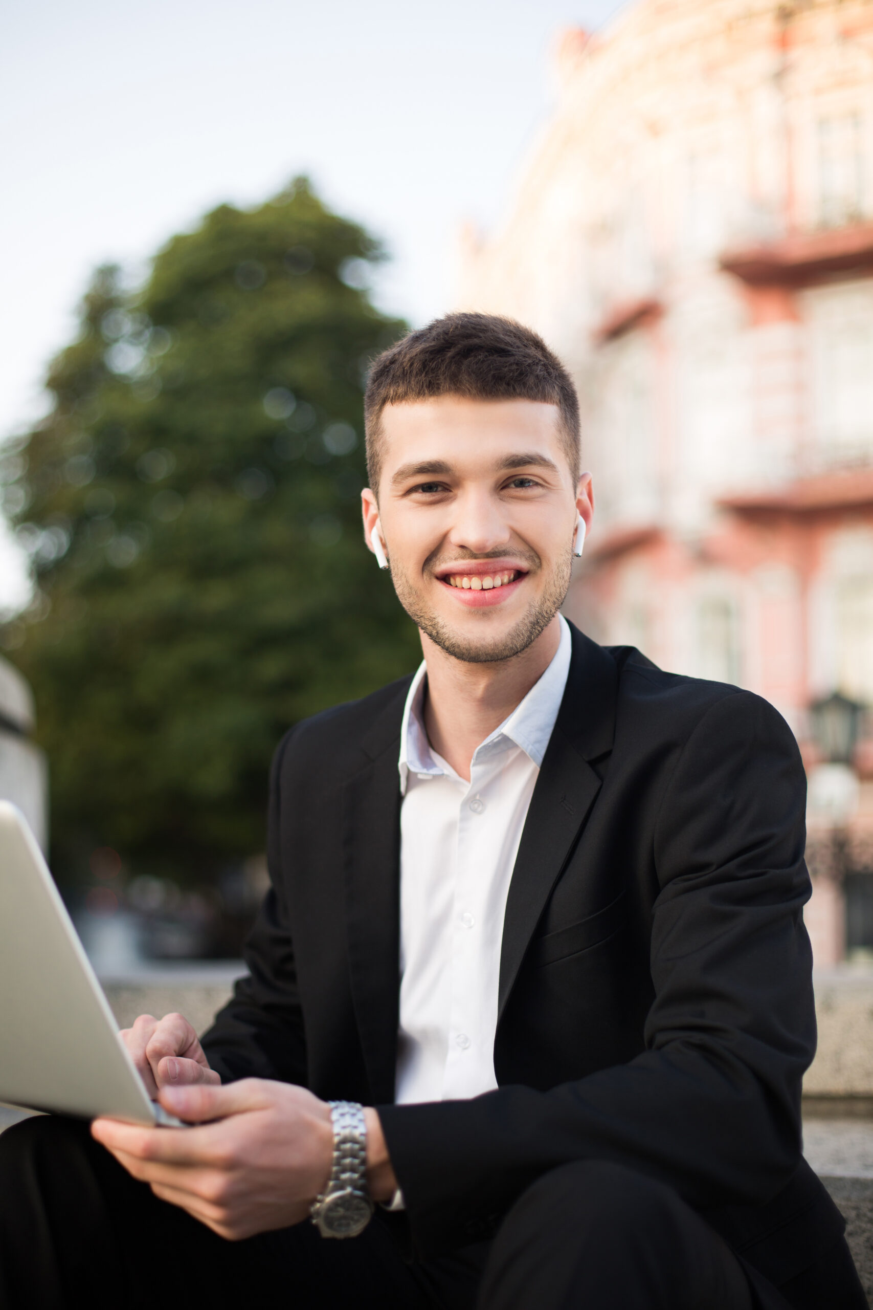 young cheerful man classic black suit white shirt with wireless earphones happily lookingin camera with laptop hands while spending time outdoor scaled